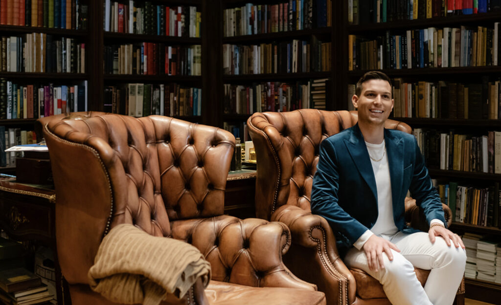 A person in a blue blazer, named Matt Fraser, sits on a brown leather chair in a library lined with bookshelves. Another similar chair is beside them.